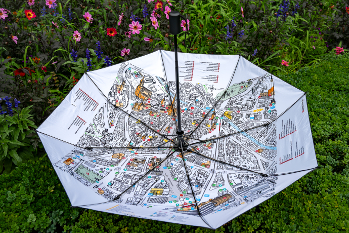 Regenschirm Innenansicht (©Markus Cadosch)  Ein aufgespannter Regenschirm liegt geöffnet im Grünen. Auf der Innenseite ist ein farbenfroher Stadtplan von Chur abgebildet, während im Hintergrund bunte Blumen für eine lebendige Szenerie sorgen.