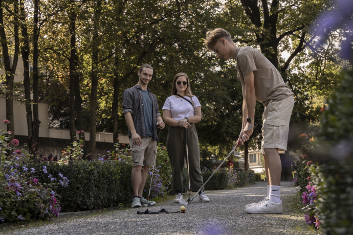 Urban Golf (© Flavio Schlegel) Three people play mini-golf in a sunlit park, surrounded by lush flowers. One is taking a shot while the others watch, creating a relaxed and fun atmosphere
