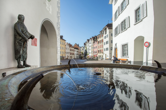 Arcas (© Yvonne Bollhalder) Historic European street with colorful buildings under a clear blue sky. Foreground features a rippling fountain near a bronze statue, creating a serene atmosphere