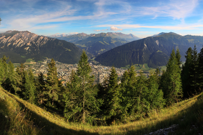 Panorama view (© Yvonne Bollhalder) Panoramic view of a lush green valley with a small town nestled among dense trees. Majestic mountains rise in the background under a blue sky