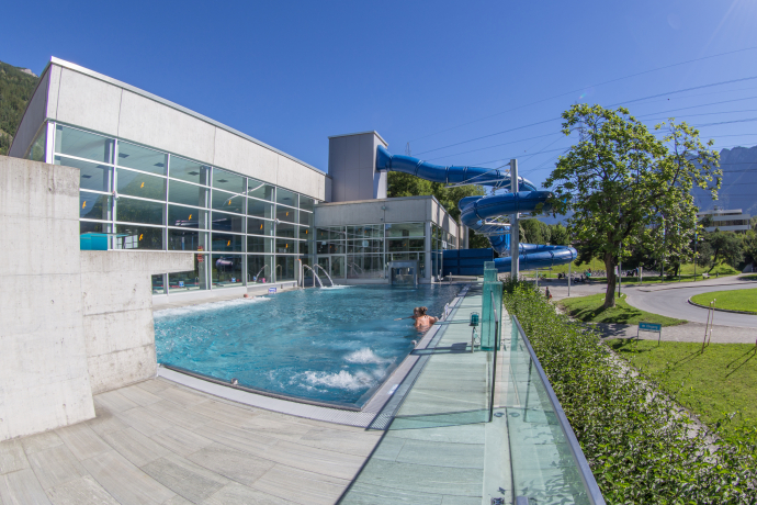 Aquamarin Obere Au (© Yvonne Bollhalder) Modern aquatic center with an outdoor pool, glass walls, and a blue waterslide. A few people relax in the sun, surrounded by greenery