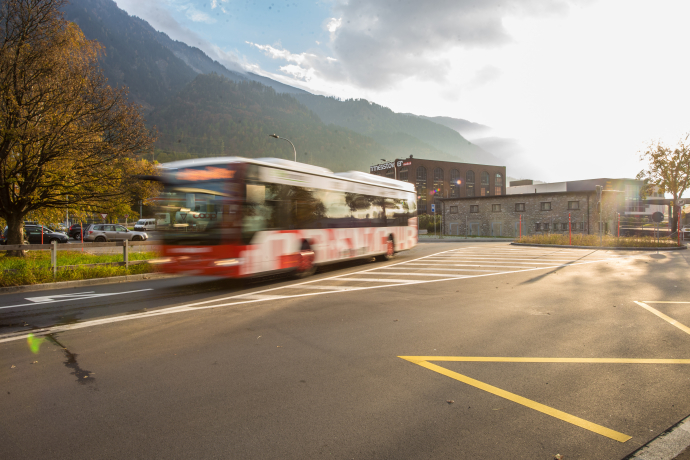 Bus Chur (© Chur Bus) A red and white bus speeds by on a quiet road with mountains and a building in the background