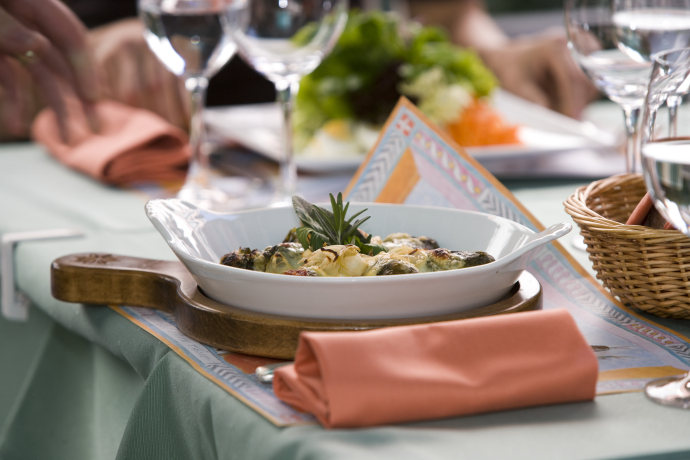 Local specialities (© Andrea Badrutt) A beautifully set table features a dish of baked escargot in a white bowl on a wooden board, accompanied by a folded orange napkin and wine glasses, conveying a gourmet dining experience