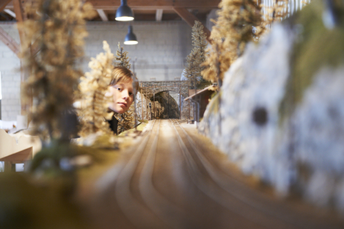 Railway museum (© railway museum) A child intently observes a detailed miniature train model with trees and a tunnel in a warmly lit room, evoking curiosity and wonder