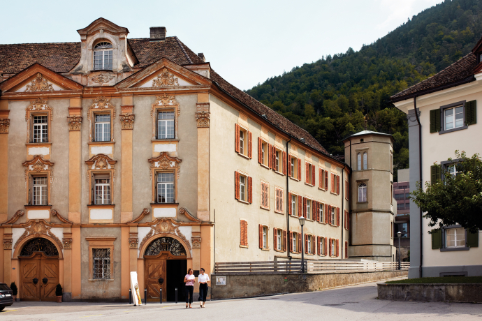 Cathedral treasury museum (©Hannes Heinzer) Historic European building with ornate architecture and orange shutters set against a forested hillside. Two people walk in front, creating a serene scene