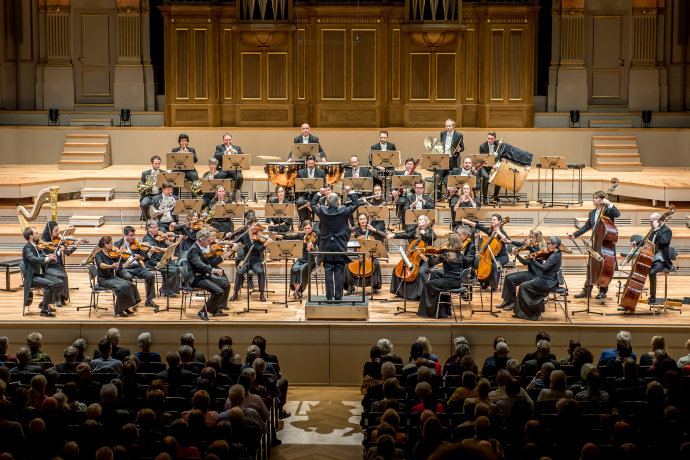 Grisons chamber philharmonic (© Grisons chamber philharmonic) An orchestra performs on a grand stage with musicians playing strings, brass, and percussion. The conductor leads passionately; audience attentively watches