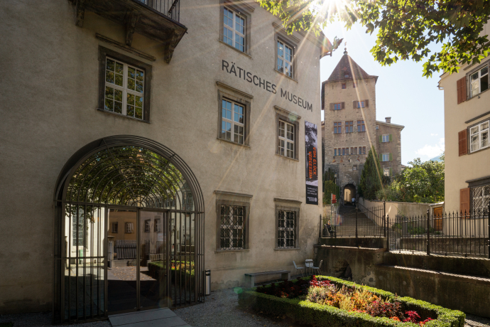 Raetian museum (© Andre Meier) Sunlit facade of Rätisches Museum with arched entrance, surrounded by manicured gardens. A historic tower rises in the background under a clear sky