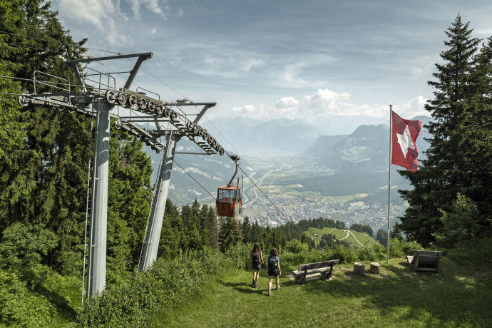 Railway Brambrüesch (© Dominik Baur) Two people walk toward a cable car in a scenic mountain landscape. A Swiss flag flutters nearby, with lush greenery and a town below