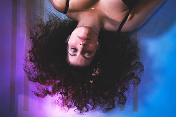 Salzkeller Chur (© Salzkeller) A woman with curly hair lies on her back in a calm, dreamy setting. Her expression is serene, surrounded by soft purple and blue lighting