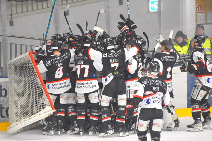 EHC Chur (© EHC) A hockey team huddles joyfully in front of a goal, raising their sticks. They wear black and white uniforms with red accents. The mood is celebratory