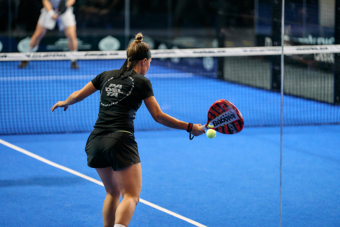 Padelta Chur (© Padelta) A woman in athletic wear prepares to hit a ball with a paddle on a bright blue padel court. A focused stance and dynamic action convey intensity