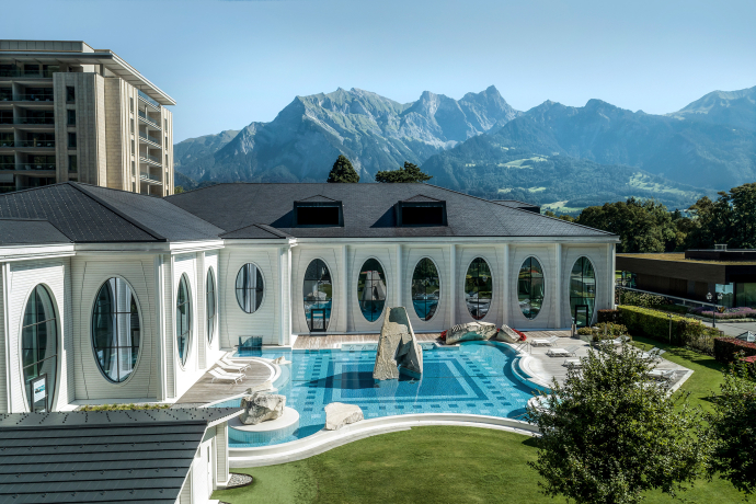 Tamina thermal bath (© Tamina thermal bath) A luxurious spa building with large oval windows overlooks a scenic mountain range. The blue pool is surrounded by lounging areas