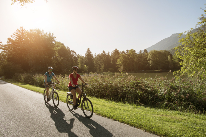 Kulinarik-Tour (© Heidiland Tourismus) Zwei Radfahrerinnen fahren entspannt auf einem sonnendurchfluteten Weg entlang eines Sees, umgeben von Bäumen und Bergen im Hintergrund. Die Szene vermittelt sommerliche Leichtigkeit und Naturgenuss.