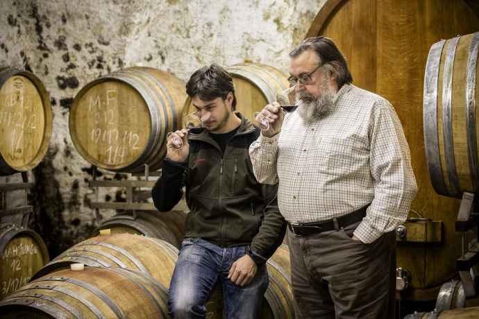 von Tscharner - Weinverkostung (© Salz&Pfeffer) Zwei Männer stehen in einem Weinkeller vor grossen Holzfässern. Sie halten jeweils ein Glas Rotwein in der Hand und prüfen konzentriert den Duft. Die Szene vermittelt Handwerkstradition und die Kunst der Weinverkostung.
